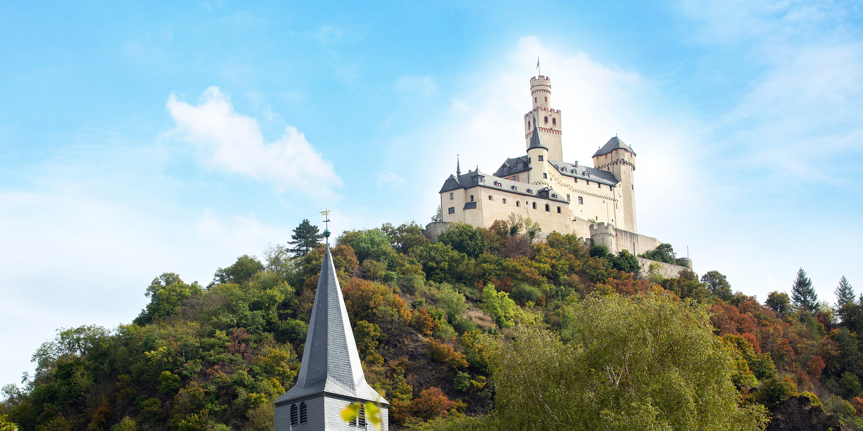 cream coloured castle on top of a hill with green foliage surrounding, and slightly cloudy blue skies