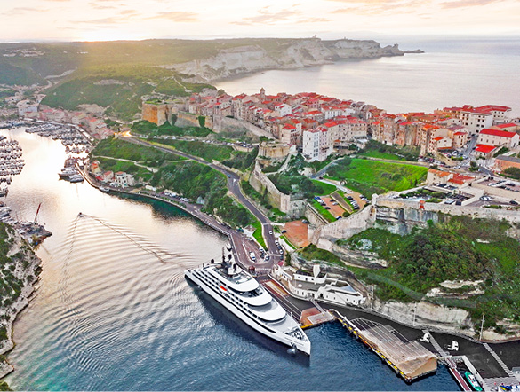 Luxury yacht docked in Mediterranean town with white buildings, near trees and the sea, as sun sets