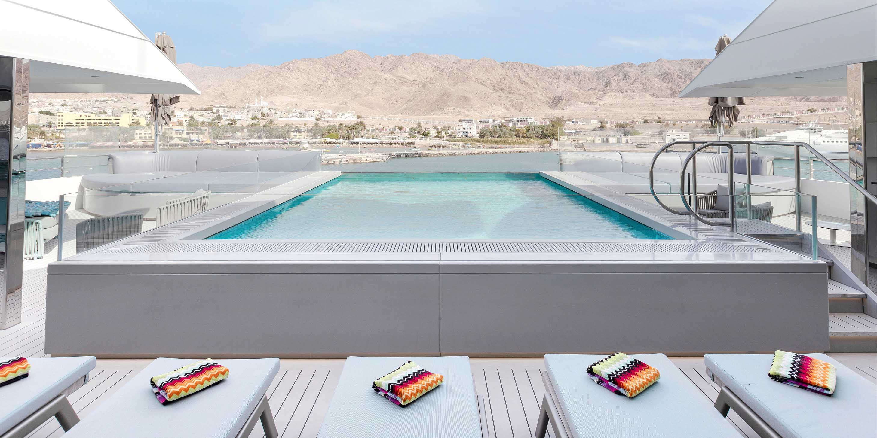 A large pool on the top deck of a yacht next to a row of sun loungers, with hills in the background